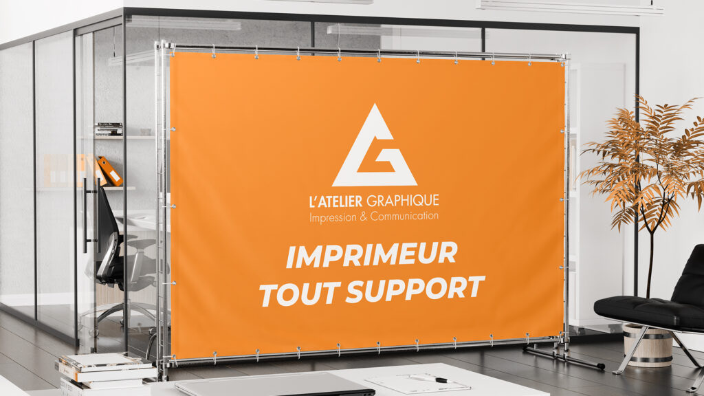 Bâche publicitaire orange grand format L'Atelier Graphique tendue sur un cadre métallique dans un bureau moderne.