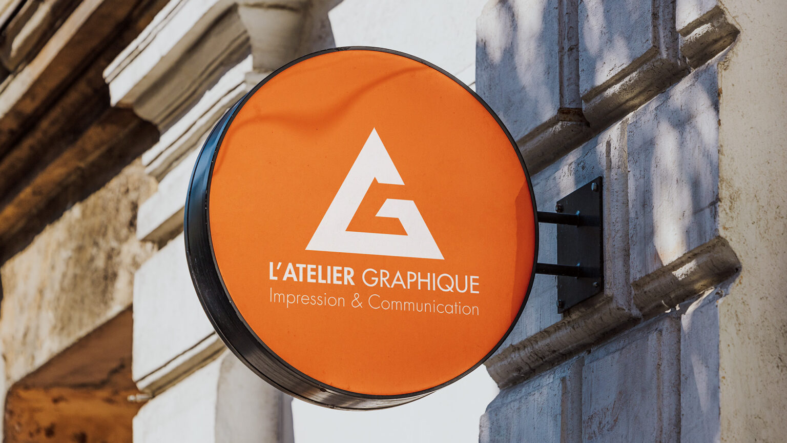 Enseigne ronde extérieure de couleur orange de L'Atelier Graphique Impression et Communication fixée sur une façade en pierre