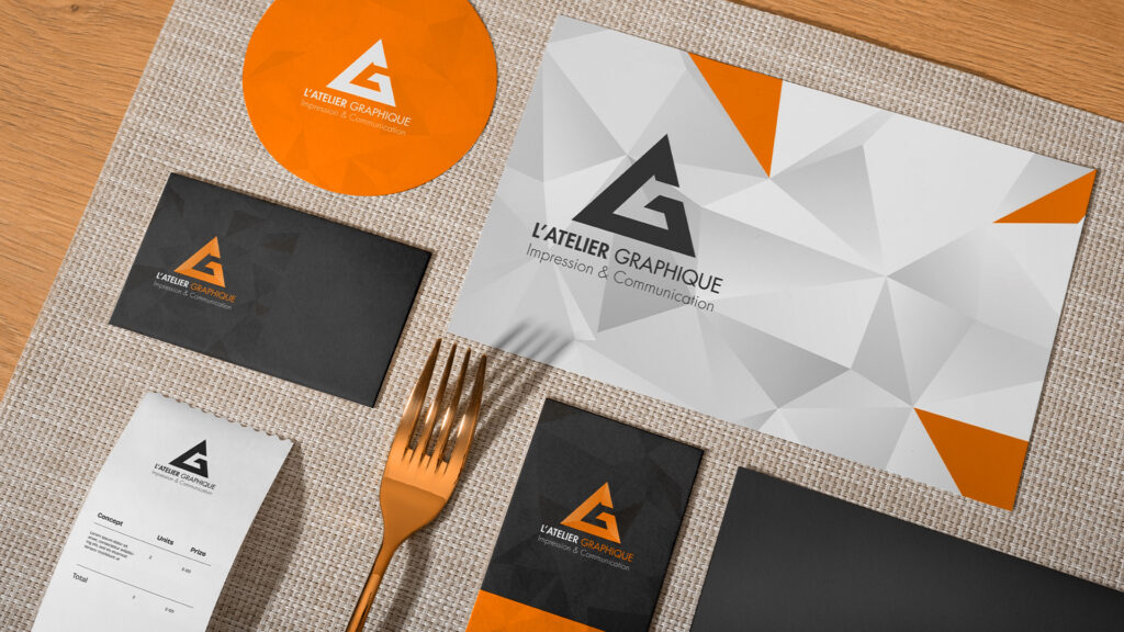 Supports imprimés pour restaurant L'Atelier Graphique comprenant un menu géométrique, un sous-verre rond orange, des cartes noires et un ticket de caisse sur un set de table avec une fourchette bronze